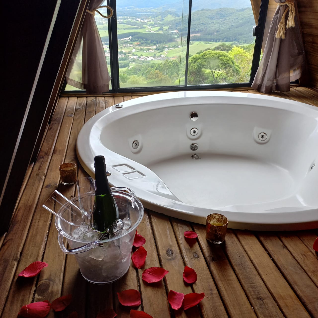 Banheira de hidromassagem junto da parede de vidro, com champanhe, pétalas e a vista do vale